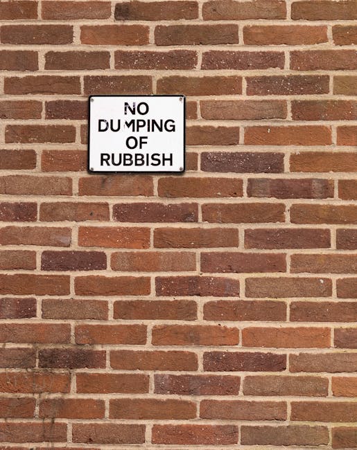 A rectangular metal sign with a white background and black text is mounted on a red brick wall. The sign reads 'NO DUMPING OF RUBBISH,' with the word 'DUMPING' partially obscured or faded, making it less legible. The brick wall features a standard running bond pattern with reddish-brown bricks and light grey mortar jointing. The bricks have a slightly textured surface and vary subtly in tone, contributing to a traditional urban setting. The lighting is diffuse, suggesting an overcast day, and there are no visible shadows or additional objects in the scene. The sign appears to serve as a reminder against illegal rubbish dumping, which aligns with the context of a rubbish removal service provider such as Rubbish Removal Putney, operating in residential or commercial areas where private waste management is necessary.